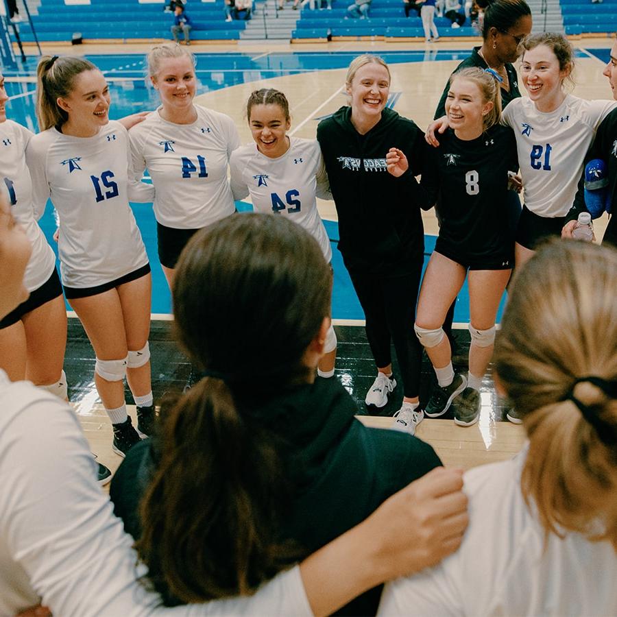 Girls volleyball players stand in circle on court smiling.